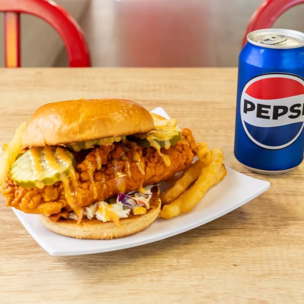 Spicy fried chicken sandwich with pickles, coleslaw, fries, and a Pepsi on a wooden table.