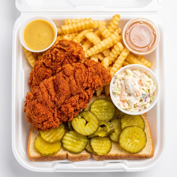 A takeout container with Nashville hot chicken tenders, crinkle-cut fries, coleslaw, pickles, and dipping sauces.